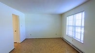 This single-floor space presents a straightforward and functional layout well-suited for a variety of uses. The room showcased in the image is finished with classic parquet wood flooring arranged in a traditional block pattern, adding a timeless warmth to the otherwise understated interior. The walls are painted in a clean, neutral off-white tone, creating a bright and open atmosphere that allows for flexible decorating possibilities.

Natural light enters the room through a window fitted with horizontal blinds, providing both illumination and privacy control. Positioned along the base of the wall beneath the window is a baseboard heating unit, indicating a reliable and evenly distributed heating system throughout the space. The ceiling is smooth and white, contributing to the room's airy feel.

Electrical outlets are distributed along the walls at standard intervals, supporting practical everyday use. The overall design is clean and minimal, with no ornate architectural detailing, making the space highly adaptable. The combination of warm wood flooring and neutral walls gives the room a neutral foundation ready to be personalized.