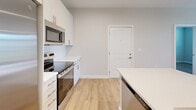 This thoughtfully designed 2-bedroom, 1-bathroom single-floor residence offers a clean, modern aesthetic throughout. Light wood-tone flooring flows consistently across all rooms, complemented by neutral warm-gray walls and crisp white trim, creating a bright and cohesive feel.

The kitchen serves as a functional centerpiece, featuring a large island with a built-in sink, white shaker-style cabinetry, white quartz countertops, a white subway tile backsplash, and a full suite of stainless steel appliances including a refrigerator with ice maker, electric range, built-in microwave, and dishwasher. Recessed lighting adds to the clean, modern finish.

The living room is generously sized and filled with natural light through large windows and a glass door with transom that opens to an outdoor area. A ceiling fan adds comfort to this welcoming space.

The primary bedroom offers a comfortable footprint with a window providing natural light and outdoor views, while the second bedroom similarly benefits from natural light and direct access to the hallway and kitchen area.

The bathroom is well-appointed with a wide vanity, light wood cabinetry, white quartz counters, a tub-shower combination with subway tile surround, and wood-look flooring.

A dedicated laundry room houses a full-size side-by-side washer and dryer with overhead shelving, adding practical convenience to the home. A separate closet provides additional storage.