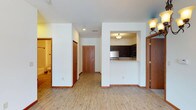 This single-floor residential space offers a clean, functional layout designed for comfortable everyday living. The main living area is open and airy, featuring light wood-style plank flooring that runs throughout, creating a warm and cohesive feel. Crisp white walls and white ceilings contribute to a bright, welcoming atmosphere, complemented by recessed and flush-mount ceiling lighting.

A defining feature of the space is the pass-through kitchen, framed by a half-wall cutout that maintains visual connectivity between the kitchen and the main living area. The kitchen is appointed with dark espresso-toned cabinetry, stainless steel appliances, and a light countertop, offering a stylish contrast against the surrounding white walls.

Rich, warm-toned wood doors are found throughout the unit, adding a consistent design element that ties the space together. A hallway leads to additional rooms, including a bathroom visible in the background, which features coordinating wood-toned finishes and tile flooring. The entry door, finished in the same warm wood tone, anchors the layout and provides a natural focal point at the end of the main corridor.

Overall, the space presents a tidy, move-in-ready environment with a practical floor plan and thoughtful design details throughout.