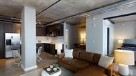 This single-floor space showcases a distinct industrial aesthetic defined by its exposed concrete ceiling and visible silver ductwork running overhead, lending the interior a raw, urban character. The open-concept layout seamlessly connects the living, kitchen, and dining areas, creating a cohesive and functional flow throughout.

The living area is anchored by a rich brown leather sofa paired with a round white coffee table, offering a warm contrast to the industrial overhead elements. Hardwood-style flooring runs throughout, adding warmth to the otherwise utilitarian design.

The kitchen features dark espresso cabinetry with a light-toned tile backsplash, stainless steel appliances, and a clean white countertop. Track lighting enhances both functionality and ambiance in this area.

Adjacent to the kitchen, a dedicated dining space is furnished with a dark table and matching chairs, illuminated by a warm-toned lamp. Structural columns punctuate the open floor plan, reinforcing the building's architectural character. The combination of industrial materials and warm furnishings creates a balanced, contemporary living environment.