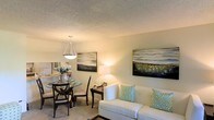 This single-floor residence offers a welcoming and well-appointed layout designed for comfortable everyday living. A hallway entry with a interior door leads into the main living area, where soft carpeted flooring and a textured ceiling create a warm, cozy atmosphere. The living room is generously sized, featuring a large window dressed with patterned drapes that invite an abundance of natural light. A decorative sunburst mirror and coordinating wall art add stylish accents to the neutral-toned walls, lending the space a polished, curated feel. The open-plan layout connects the living room seamlessly to the dining area, which is anchored by a round glass-top dining table with dark wood chairs and illuminated by a modern pendant light overhead. Large-scale landscape artwork adorns the walls, reinforcing the cohesive, contemporary aesthetic throughout. A glimpse of the kitchen is visible beyond the dining space, suggesting a practical and connected flow between living and cooking areas. The overall ambiance is light, airy, and inviting, with a neutral color palette and thoughtful décor that creates a harmonious and comfortable environment throughout the home.