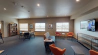 The Family Room is a well-appointed, single-floor community space designed to accommodate a variety of activities and social gatherings. The layout is thoughtfully divided into several functional zones, each contributing to the room's versatile character.

The lounge area serves as the focal point of the space, furnished with bold orange accent chairs, a striped sofa, and low-profile side tables arranged around a wall-mounted flat-screen television. An open wire media console anchors this zone, adding an industrial touch to the otherwise warm, neutral palette. Recessed lighting illuminates the carpeted floor throughout this section.

Adjacent to the lounge, a more intimate seating arrangement features paired orange chairs flanking a small table, positioned near multiple windows fitted with horizontal blinds. These windows allow generous natural light to fill the space and offer views of the surrounding neighborhood. Framed artwork decorates the walls, adding color and visual interest.

A separate dining and meeting area transitions into a checkerboard-tiled floor, furnished with a dark conference-style table and task chairs, suitable for group activities or meetings.

The fully equipped kitchen rounds out the space, featuring dark wood cabinetry, stainless steel appliances including a refrigerator, range, dishwasher, and microwave, along with durable wood-look flooring and ample counter space for food preparation and entertaining.
