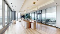 Located at 1950 Broadway in Oakland, CA, this sophisticated single-floor space combines contemporary design with stunning elevated views of the Oakland skyline and beyond. The living room is the centerpiece of the space, wrapped in expansive floor-to-ceiling glass curtain walls that flood the interior with natural light and frame breathtaking panoramic views of the city and surrounding landscape. Light oak hardwood flooring runs throughout, complementing the bright, airy atmosphere. A statement dining table with a rich wood base is surrounded by curved cream upholstered chairs beneath a cascading arrangement of circular gold-accented pendant lights, adding an elegant focal point to the open layout. A seating area near the glass perimeter features plush, rounded accent chairs positioned to take full advantage of the sweeping views, anchored by sleek structural columns. The kitchen is a well-appointed galley-style space finished with warm wood cabinetry, dark lower cabinets, black granite countertops, and a striking hexagonal mosaic tile backsplash. Stainless steel appliances, including a built-in oven, induction cooktop, and full-size refrigerator, provide comprehensive functionality. The overall ambiance of the space is refined and contemporary, blending high-end finishes with an abundance of natural light and commanding urban views.