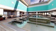 Located in the heart of downtown Dallas at 350 N Ervay St, this rooftop amenity deck offers an exceptional outdoor retreat defined by modern design and urban sophistication. The space is anchored by a stunning resort-style pool finished with gleaming mosaic tile, surrounded by a broad wood-plank deck lined with cushioned chaise lounges and low-profile side tables — ideal for sunbathing with panoramic city skyline views as the backdrop. The building's bold teal and white exterior panels frame the deck, lending the space a vibrant, contemporary aesthetic. A striking floor-to-ceiling travertine stone fireplace, detailed with iridescent mosaic tile accents and lava rock at its base, serves as a focal point in the covered lounge area, providing warmth and ambiance during cooler evenings. Ceiling fans overhead keep the covered sections comfortable year-round. An outdoor kitchen and bar area, complete with a stainless steel range hood, bar-height seating, and dining tables, make this deck well-suited for entertaining. Potted tropical plants add natural greenery throughout, softening the urban setting. Whether relaxing poolside, gathering around the fireplace, or dining al fresco, this rooftop deck delivers a compelling blend of comfort, style, and downtown Dallas energy.