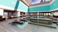 Located in the heart of downtown Dallas at 350 N Ervay St, this rooftop amenity deck offers an exceptional outdoor retreat defined by modern design and urban sophistication. The space is anchored by a stunning resort-style pool finished with gleaming mosaic tile, surrounded by a broad wood-plank deck lined with cushioned chaise lounges and low-profile side tables — ideal for sunbathing with panoramic city skyline views as the backdrop. The building's bold teal and white exterior panels frame the deck, lending the space a vibrant, contemporary aesthetic. A striking floor-to-ceiling travertine stone fireplace, detailed with iridescent mosaic tile accents and lava rock at its base, serves as a focal point in the covered lounge area, providing warmth and ambiance during cooler evenings. Ceiling fans overhead keep the covered sections comfortable year-round. An outdoor kitchen and bar area, complete with a stainless steel range hood, bar-height seating, and dining tables, make this deck well-suited for entertaining. Potted tropical plants add natural greenery throughout, softening the urban setting. Whether relaxing poolside, gathering around the fireplace, or dining al fresco, this rooftop deck delivers a compelling blend of comfort, style, and downtown Dallas energy.