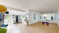 Located at Design Place in Miami's vibrant MiMo district, this fitness facility offers a thoughtfully designed workout environment spread across a single, open floor. The gym is bathed in natural light, courtesy of expansive floor-to-ceiling windows and clerestory glazing that illuminate the light wood flooring and crisp white walls throughout.

The cardio zone features a row of treadmills and elliptical machines positioned to face large glass doors that open directly to an outdoor pool area, allowing members to enjoy tropical views and fresh air while exercising. Mounted flat-screen TVs provide additional entertainment. A bold, colorful mural depicting athletic silhouettes energizes the space, adding a dynamic visual focal point to the cardio section.

The strength training area is equipped with a multi-station cable machine and various resistance equipment, complemented by full-length wall mirrors that enhance the sense of openness. A dedicated functional fitness zone offers ample open floor space for stretching, yoga, or step aerobics, supported by a rack stocked with stability balls, medicine balls, and resistance accessories.

The facility also includes a water fountain station and restroom access. Motivational typography displayed on the walls reinforces the gym's active atmosphere. The overall design balances vibrant artistic elements with a clean, functional layout, creating an inviting space for a comprehensive fitness experience.