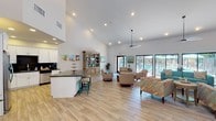 Located at 1055 W Baseline Rd in Mesa, Arizona, this single-floor community clubhouse offers an expansive, open-concept interior designed for gathering and relaxation. Soaring vaulted ceilings amplify the sense of space throughout, while recessed lighting and large sliding glass doors allow abundant natural light to fill the interior. Light wood-look tile flooring runs seamlessly across the entire space, lending a clean, modern aesthetic.

The well-appointed kitchen features crisp white cabinetry, dark granite countertops, a dark tile backsplash, stainless steel appliances including a full-size refrigerator, and a spacious island with bar seating. Colorful framed artwork lines the adjacent wall, adding a vibrant touch to the area.

The open lounge area is furnished with multiple seating arrangements, including a teal sofa, patterned accent chairs, and a rustic wooden high-top table with industrial-style stools. A wall-mounted flat-screen TV and a decoratively distressed media console serve as focal points for the entertainment zone. Ceiling fans contribute to comfort throughout.

Large sliding glass doors and floor-to-ceiling windows along one wall provide direct views of and access to the outdoor pool area, seamlessly connecting the interior with the exterior amenity space. The overall ambiance is bright, welcoming, and thoughtfully designed for community use.
