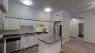 This single-floor residence is designed with a practical and cohesive layout that connects the kitchen and living spaces seamlessly. The kitchen is a standout feature, outfitted with white shaker-style cabinetry, dark granite countertops, and a classic subway tile backsplash. A stainless steel refrigerator and built-in microwave provide modern functionality, while the double-basin sink sits beneath under-cabinet range ventilation. A freestanding center island with matching granite countertops offers additional prep space and serves as a natural dividing point between the kitchen and the rest of the unit. Flush-mount ceiling lights illuminate the kitchen area, and wood-look flooring adds warmth and durability underfoot.

The living area transitions to plush carpeting and features vaulted ceilings with crown molding detail. A ceiling fan with a light fixture provides both comfort and ambient lighting. Large windows along the far wall allow generous natural light to fill the room, with views of surrounding greenery and access to what appears to be a private balcony or patio. Track lighting and neutral wall tones complete the inviting, move-in-ready atmosphere throughout the space.