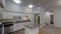 This single-floor residence is designed with a practical and cohesive layout that connects the kitchen and living spaces seamlessly. The kitchen is a standout feature, outfitted with white shaker-style cabinetry, dark granite countertops, and a classic subway tile backsplash. A stainless steel refrigerator and built-in microwave provide modern functionality, while the double-basin sink sits beneath under-cabinet range ventilation. A freestanding center island with matching granite countertops offers additional prep space and serves as a natural dividing point between the kitchen and the rest of the unit. Flush-mount ceiling lights illuminate the kitchen area, and wood-look flooring adds warmth and durability underfoot.

The living area transitions to plush carpeting and features vaulted ceilings with crown molding detail. A ceiling fan with a light fixture provides both comfort and ambient lighting. Large windows along the far wall allow generous natural light to fill the room, with views of surrounding greenery and access to what appears to be a private balcony or patio. Track lighting and neutral wall tones complete the inviting, move-in-ready atmosphere throughout the space.