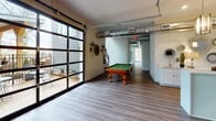 This single-floor space offers a versatile, open-concept layout designed for both relaxation and recreation. The interior is finished with durable wood-look plank flooring throughout, complemented by clean white and soft neutral walls that create a bright, welcoming atmosphere. A dedicated lounge area anchors one end of the space, furnished with a gray upholstered sofa, mid-century style armchairs, a low-profile coffee table, and layered area rugs. Pendant lighting with brass-toned fixtures hangs above, while decorative wall-mounted sconces and an armillary sphere accent light add distinctive character. A wall-mounted flat-screen television is positioned for comfortable viewing from the seating area. Adjacent to the lounge, a full-size pool table with ornate wooden legs serves as a standout recreational feature. A striking floor-to-ceiling glass panel garage door opens the space to an outdoor patio, flooding the interior with natural light and providing seamless indoor-outdoor access. Exposed ductwork along the ceiling lends an industrial edge to the otherwise polished interior. A wet bar or service counter area is visible near the back, and a corridor connects to additional areas of the building. Decorative greenery and layered accent pieces complete the modern, curated aesthetic.