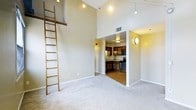 This thoughtfully designed single-floor residence makes a memorable first impression with its dramatically high ceilings that lend an open, airy quality to the main living area. A standout architectural feature is the tall wooden ladder mounted to the wall, hinting at the vertical drama the elevated ceiling height provides. The living space is finished with neutral-toned carpet and warm beige walls, creating a calm and inviting atmosphere throughout.

Pendant lighting and track fixtures are suspended from the soaring ceiling, casting a warm ambient glow across the space. The open layout allows for flexible furniture arrangements and a comfortable sense of flow between areas.

Adjacent to the main living area, the kitchen is visible through a transitional opening and is equipped with stainless steel appliances, including a range and refrigerator. Rich, dark wood cabinetry lines the walls and provides ample storage, while a countertop workspace offers functional prep space. A circular wall sconce adds a decorative accent to the kitchen area.

Overall, the space balances practicality with character — the high ceilings and architectural ladder detail give it a loft-like quality, while the warm finishes and integrated kitchen ensure everyday comfort and functionality.
