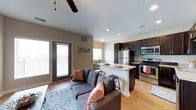 This single-floor space showcases a thoughtfully designed open-concept layout that effortlessly connects the living, dining, and kitchen areas into one cohesive living environment. The living area is anchored by warm hardwood-style flooring and features a ceiling fan overhead, while large windows and a glass-paneled door flood the space with abundant natural light and provide access to the outdoors.

The kitchen is a standout feature, boasting rich dark cabinetry that creates a striking contrast against the light-toned granite countertops. A raised breakfast bar with seating serves as both a functional prep space and a casual dining spot. Stainless steel appliances, including a refrigerator, range, and microwave, lend a modern, polished look to the culinary space.

Adjacent to the kitchen, a compact dining nook sits beneath a window, offering a cozy spot for meals. The living area flows naturally from the kitchen, creating an inviting atmosphere ideal for both relaxing and entertaining. Neutral wall tones complement the dark cabinetry and light flooring, giving the space a warm, balanced aesthetic. Recessed lighting and textured ceilings add subtle architectural character throughout. Overall, the space combines practicality with modern design sensibilities.
