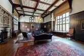 Located in Washington, DC's Dupont Circle neighborhood, this distinguished historic space exudes old-world grandeur across a generous single floor. The architecture is defined by deep-toned dark wood wall paneling throughout, intricate carved millwork, and leaded glass windows that cast soft, filtered natural light into each room.

The sweeping hallway sets an immediate tone of elegance, featuring an elaborately detailed plaster ceiling with interlacing relief patterns and warm hardwood floors layered with fine area rugs. A dramatic great hall commands attention with soaring beamed ceilings, a brass chandelier, tall leaded glass windows, a fireplace, and a grand piano — evoking a Tudor Revival aesthetic.

The library is a refined retreat lined floor-to-ceiling with built-in dark wood bookshelves filled with volumes, complemented by a decorative fireplace, exposed beam ceiling details, and comfortable seating arranged around a central coffee table. A formal conference or meeting room features a large oval wooden table, leather-padded chairs, mica-shade lamps, and richly paneled walls adorned with oil paintings. Additional rooms display carved pilasters, antique furnishings, and curated artwork throughout. The space as a whole conveys an atmosphere of scholarly sophistication and historic institutional character.