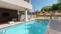 This outdoor pool area presents a well-appointed retreat defined by clean, contemporary design. The centerpiece is a generously sized rectangular pool with clear turquoise water, accessible via built-in steps and stainless steel handrails. Depth markers are visible along the pool's edge, and the surrounding deck is finished in smooth, light-toned concrete that extends comfortably around all sides.

A row of modern white lounge chairs lines the pool deck, several accented with patterned throw pillows, offering ample space for sunbathing. Bright yellow umbrellas punctuate the perimeter, providing shade over additional seating areas, including dining-height tables and chairs positioned along the far end.

Patches of artificial turf add visual contrast against the neutral hardscape, creating designated lounge zones. The space is enclosed by horizontal slatted privacy fencing, with mature trees visible beyond the perimeter, contributing a natural backdrop. A covered section along the building edge features brick detailing, large windows, and a wall-mounted television, extending the usability of the space. The overall ambiance is bright, open, and well-suited for outdoor relaxation.