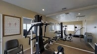 This dedicated fitness room offers a functional and well-appointed space for strength training and exercise. The room is outfitted with multiple cable and weight machines, providing a comprehensive workout experience without leaving the premises. Large wall-to-wall mirrors span one side of the room, visually expanding the space and allowing users to monitor their form during exercise sessions. The carpeted flooring adds a degree of comfort underfoot, while recessed lighting and multiple windows with blinds ensure the space remains well-lit throughout the day. Natural light filters in through the windows, creating a bright and energizing atmosphere. The textured ceiling adds a subtle architectural detail, and the neutral warm-toned walls contribute to a clean, focused environment. Ventilation is provided via overhead vents, keeping the space comfortable during use. Overall, this fitness room is thoughtfully designed to serve as a practical and motivating space for residents or occupants looking to maintain an active lifestyle on-site.