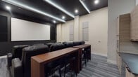 This purpose-built screening room is thoughtfully designed to deliver an immersive cinematic experience. The focal point is a large projection screen mounted against a dark accent wall, flanked by acoustic panels that optimize sound quality throughout the space. A ceiling-mounted projector is positioned toward the rear of the room, ensuring a clear, unobstructed image.

Multiple rows of black leather recliner seats are arranged in a theater-style layout, each equipped with built-in cup holders and tray tables for added convenience. The seating faces the screen directly, maximizing sightlines for all viewers. Along the side and rear walls, additional acoustic panels are strategically placed to further enhance sound performance and reduce unwanted reverberation.

The ceiling features a dramatic dark finish with recessed lighting and integrated LED strip lighting that provides ambient illumination without interfering with the viewing experience. Patterned gray carpet tile covers the floor throughout, contributing to the room's acoustic character.

A built-in kitchenette with wood cabinetry and a countertop is situated at the rear of the room, accompanied by a wooden bar-height counter and black bar stools, offering a casual seating and refreshment area. An illuminated emergency exit door is also present, ensuring safety compliance.