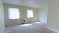 This single-floor space features a spacious, open main room finished in a clean, neutral palette of soft cream and off-white tones. Wall-to-wall carpeting in a light beige shade covers the floor, contributing to a quiet and comfortable atmosphere. Two windows fitted with white horizontal blinds allow generous amounts of natural light to fill the room, casting bright patches across the floor and enhancing the overall sense of openness. A wall-mounted window air conditioning unit is positioned beneath one of the windows, providing climate control. The walls are smooth and painted in a light tone that complements the carpeting, while the textured popcorn ceiling adds a classic touch characteristic of traditional construction. Paneling detail is visible along a portion of one wall, adding a subtle architectural element to the otherwise straightforward design. The room is currently unfurnished, allowing the full scale and layout of the space to be appreciated. The overall ambiance is bright, clean, and functional, offering a blank canvas suitable for a variety of uses.