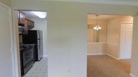 This single-floor residence offers a practical and comfortable layout with distinct living zones that flow naturally into one another. The kitchen is outfitted with dark cabinetry, stainless steel appliances including a refrigerator and stove, and easy-to-maintain tile flooring. A skylight overhead bathes the kitchen in natural light, enhancing its functional character. The kitchen opens directly to the main living area, which features neutral wall tones and plush carpeting that adds warmth underfoot. Crown molding runs throughout the space, lending a refined architectural touch to the otherwise clean and understated design. Adjacent to the living area, a dedicated dining space is highlighted by a classic chandelier that serves as a central focal point, providing warm ambient lighting for gatherings. The dining room continues the neutral color palette and carpeted flooring, maintaining a cohesive aesthetic across the home. White base trim and panel-style doors add to the traditional character of the interior. The open connectivity between the kitchen, living, and dining areas makes the home feel welcoming and easy to navigate, while the consistent finishes and thoughtful lighting create a unified and livable environment throughout the single floor.