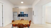 Located at 626 NW 11th Ave in Miami, FL, this efficiently designed 3-bedroom, 2-bathroom unit offers a clean, modern aesthetic across a single floor. The open-plan kitchen and living area serves as the heart of the space, featuring light hardwood-style flooring, white walls, and abundant natural light from windows overlooking the surroundings. The kitchen is outfitted with sleek, light wood cabinetry extending floor to ceiling, a dark tile backsplash, stainless steel appliances including a full-size refrigerator, over-range microwave, and dishwasher. A wooden dining table with amber acrylic chairs anchors the dining area, while the living space accommodates a wall-mounted TV and comfortable seating. Each of the three bedrooms maintains the same minimalist aesthetic with white walls, light flooring, and individual windows that bring in natural light. The bedrooms include built-in wardrobe storage and dedicated desk areas, making them functional for both rest and work. The two bathrooms are finished with large-format neutral tiles; one features a walk-in shower with floor-to-ceiling stone-toned tile surround, while the other offers a separate shower area. The overall design emphasizes clean lines, neutral tones, and practical use of space throughout.