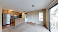 This single-floor space offers an open-concept layout that seamlessly connects the kitchen and living area, creating a functional and cohesive environment. The kitchen is equipped with warm honey-toned wood cabinetry, stainless steel appliances, and a built-in microwave. A peninsula-style breakfast bar with a smooth countertop extends into the living space, providing both additional workspace and a casual dining option. A tiled backsplash adds a clean, finished look to the kitchen area.

The transition from the kitchen to the living area is defined by a shift from tile to plush carpet flooring, with a decorative brick tile section near the entryway adding a touch of character. The living area is generously sized, featuring neutral beige walls and recessed lighting that contribute to a warm, inviting ambiance. A window along the exterior wall allows natural light to filter in, brightening the space.

Near the entryway, a stacked washer and dryer unit is conveniently integrated into the layout, offering practical in-unit laundry access. The front door area features durable tile flooring, ideal for a transitional entry zone. Overall, the space is designed for comfortable, everyday living with thoughtful attention to functionality.
