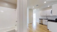 Musical String House is a single-floor residence that combines a clean, modern aesthetic with functional design. The space is characterized by crisp white walls and light-toned hardwood flooring that runs throughout, reflecting natural light and lending a sense of openness to the interior.

The kitchen is equipped with white shaker-style cabinetry, a dark contrasting countertop, and stainless steel appliances, including a dishwasher. The cabinetry extends to the upper walls, offering ample storage. Recessed lighting overhead provides consistent illumination across the space.

An open corridor connects the kitchen to the main living area, which benefits from a large window that draws in natural light, creating an airy and welcoming atmosphere. The layout promotes a seamless flow between spaces.

The bathroom is positioned adjacent to the main living areas and features a full bathtub with an overhead shower fixture, set against clean white surroundings. The overall design throughout the residence prioritizes simplicity and practicality, making it a well-appointed, move-in-ready space with a bright, neutral palette.