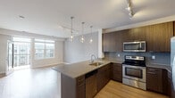 This contemporary single-floor residence showcases a thoughtfully designed open-concept layout that seamlessly connects the kitchen and living areas. The kitchen is a standout feature, boasting rich dark wood cabinetry, a gray tile backsplash, and a full suite of stainless steel appliances including a French door refrigerator, electric range with microwave above, and dishwasher. A peninsula with a quartz-style countertop and undermount sink serves as both a functional workspace and a visual divider between the kitchen and the living area, elegantly illuminated by a series of modern teardrop pendant lights overhead. Track lighting throughout the kitchen enhances the sleek, modern aesthetic. Light-toned hardwood flooring runs through the main living and kitchen spaces, contributing to a bright and airy ambiance, further complemented by a large window that floods the area with natural light. The bedroom is generously sized, featuring soft neutral carpeting, soft gray walls, and a well-placed window offering an exterior view. Adjacent to the bedroom is a full bathroom with dual-sink vanity, dark wood cabinetry with quartz countertops, a large mirror, tile flooring, and a combined tub-shower unit. The overall design palette of warm wood tones, neutral walls, and modern finishes creates a cohesive and inviting living environment.