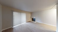 This single-floor residence offers a straightforward and functional layout designed for comfortable everyday living. The living room serves as the focal point of the home, featuring light hardwood-style flooring that runs throughout the main areas and a classic tile-surround fireplace with a white mantel and brass-accented glass doors. A large vertical-blind window treatment allows natural light to fill the space while offering privacy and light control, contributing to the room's bright, neutral ambiance. The walls are finished in soft, warm neutral tones that complement the clean aesthetic throughout.

The kitchen is efficiently arranged in an L-shaped layout, outfitted with white cabinetry, granite-style laminate countertops, a double stainless steel sink, and white appliances including an electric range and dishwasher. A window above the sink introduces additional natural light into the space.

The bedroom is generously proportioned, finished with plush carpeting and a window fitted with horizontal blinds, along with a baseboard heater for climate control.

The bathroom is clean and practical, featuring a white vanity with cabinet storage, a granite-style countertop, a wall mirror, and light hardwood-style flooring that ties the space together cohesively. Overall, this residence presents a tidy, move-in-ready environment with a consistent and understated design palette throughout.