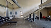 Located at 4915 Misty Oaks Dr in Charlotte, NC, this two-story fitness center offers a comprehensive workout facility designed to accommodate a wide range of exercise needs. The main floor is expansive and features patterned carpet flooring with herringbone-style tile accents, soaring ceilings, and large floor-to-ceiling mirrors that amplify the sense of space. The ground level is outfitted with an extensive selection of strength and cardio equipment, including cable machines, multi-station weight systems, free weight racks with dumbbells and kettlebells, adjustable benches, and spin bikes. A staircase connects the main floor to the upper-level mezzanine, which is enclosed by a sleek cable-railing system. The mezzanine provides additional open workout space with a wall-mounted flat-screen TV, stability balls, and windows that bring in natural light and views of the surrounding greenery. A dedicated cardio room on the upper level houses treadmills, ellipticals, and other cardio machines beneath a ceiling fan, with a window overlooking mature trees. The facility is equipped with built-in speakers, climate control, and ample overhead lighting throughout, creating a functional and well-maintained environment for fitness.