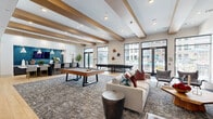 Located at 7800 E Peakview Ave in Centennial, Colorado, this single-floor community living space offers a generously sized, open-plan layout designed for both socializing and relaxation. The room is anchored by striking exposed wood beams that run across the ceiling, adding warmth and architectural character to the bright, modern interior.

The space is organized into several distinct activity zones. A central lounge area features a plush sectional sofa and accent chairs arranged around a patterned area rug, complemented by a bold textured fireplace wall clad in geometric gray tile, which also hosts a wall-mounted flat-screen television. Adjacent to the lounge sits a full-size billiards table, serving as a natural social focal point.

A dedicated dining and entertainment area is furnished with bar-height seating arranged in front of a deep teal accent wall, pendant lighting, and a large TV, creating an ideal setting for gatherings. A separate booth-style seating nook along a gray accent wall provides an additional casual dining or meeting area.

The entrance lobby features a tufted bench, vibrant abstract artwork, and the same distinctive textured tile wall, setting an upscale tone. Floor-to-ceiling windows and glass double doors flood the interior with natural light while offering direct views and access to an outdoor pool and patio area. Light hardwood flooring throughout ties the entire space together with a clean, contemporary aesthetic.