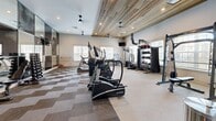 Located on Welaunee Boulevard in Tallahassee, Florida, this fitness center offers a thoughtfully designed workout space across a single floor. The room makes a strong architectural impression with its striking slatted wood ceiling, which adds warmth and texture while complementing the clean, modern aesthetic of the white walls and neutral-toned carpet flooring laid in a geometric pattern.

A full wall of large mirrored panels visually expands the space and reflects the full range of equipment throughout the room. Multiple windows fitted with white blinds line the walls, inviting ample natural light and providing views of the surrounding exterior. Ceiling fans and recessed lighting ensure the environment remains comfortable and well-lit during any workout.

The fitness center is comprehensively outfitted with a wide variety of equipment, including treadmills, elliptical trainers, a stair climber, a rowing machine, and weight benches. Strength training is well supported with a cable machine, free weights, kettlebells, and medicine balls organized on a dedicated rack. Wall-mounted televisions provide entertainment throughout the space. Safety equipment, including a fire extinguisher, is also present. The overall ambiance is modern and functional, designed to support a full range of cardio and strength training activities.