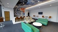 This single-floor lounge is a visually striking space defined by its bold artistic elements and contemporary design. The most commanding feature is the large-scale mural adorning an entire accent wall, rendered in vivid blues, whites, and bursts of warm color, depicting an intricate, flowing composition. Complementing this, the ceiling continues the artistic theme with a detailed painted floral and botanical motif, framed by indirect cove lighting and recessed spotlights that illuminate the space evenly.

The layout is divided into distinct zones. A bar counter with wood-paneled detailing and counter-height stools provides a casual dining or beverage area, accompanied by a mini-fridge and microwave. The central seating area features mid-century-inspired lounge chairs in neutral tones paired with a round white coffee table, while vibrant teal sectional sofas add a pop of color throughout. High-top tables with sleek chairs offer additional seating options along the perimeter. A wall-mounted flat-screen television is positioned for easy viewing from multiple seating areas. The flooring transitions between light tile and charcoal carpet, further defining each functional zone within the space.