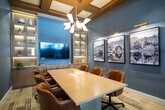 Located at 303 Arrowhead Lane in South Setauket, NY, this refined conference room offers a polished and professional environment designed for productive meetings. The centerpiece of the room is a generously sized natural wood conference table surrounded by plush cognac leather armchairs, providing both comfort and style for attendees. A large flat-panel display is mounted prominently at one end of the room, flanked by illuminated built-in shelving units that add warmth and a curated aesthetic with decorative accents. The opposite wall features a striking series of large-format black-and-white landscape photographs, each individually lit by sleek gold wall sconces that enhance the room's upscale ambiance. Overhead, a multi-arm chandelier with globe shades serves as an elegant focal point, complemented by a coffered wood ceiling detail that adds architectural character. The room is finished with textured gray wall coverings and a patterned carpet that grounds the space. Floor-to-ceiling steel-framed glass walls on one side allow visibility into the adjacent corridor and fitness area, bringing in natural light while maintaining a sense of openness. Integrated cable management in the table ensures a clean, functional workspace.