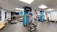 This fitness facility at 91 Valley Stream Dr in Ottawa, Ontario, offers a comprehensive workout environment spread across a single, open floor. The space is divided into two distinct zones designed to accommodate a wide range of exercise activities. The main area features a robust selection of cardio equipment, including multiple treadmills and elliptical machines, positioned alongside a striking landscape mural that adds visual energy to the room. Teal-accented structural columns and wall-mounted flat-screen TVs contribute to the modern, motivating aesthetic, while a drop ceiling with recessed panel lighting ensures the space is bright and well-illuminated throughout. A second zone is dedicated to strength training and functional fitness, housing cable machines, resistance equipment, rowing machines, stationary bikes, adjustable benches, and a rack of dumbbells and medicine balls. This area is energized by a bold motivational typography mural covering an entire wall. Rubber flooring throughout both zones provides durability and safety underfoot. Seating benches and full-length mirrors offer practical convenience for users. Large windows along one wall allow natural light to fill portions of the space, creating an open and airy atmosphere. The facility is clean, organized, and thoughtfully designed to support a full range of fitness goals.