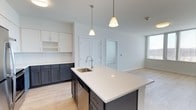 This single-floor, two-bedroom, two-bathroom residence showcases a thoughtfully designed open-concept layout that seamlessly connects the kitchen and living areas. The kitchen is a standout feature, highlighted by a large central island topped with a crisp white quartz countertop and an integrated stainless steel sink with a modern pull-down faucet. A striking two-tone cabinetry scheme pairs dark charcoal lower cabinets with bright white upper cabinets, creating a bold yet sophisticated aesthetic. A white subway tile backsplash adds texture and continuity to the design. Two cone-shaped pendant lights hang above the island, providing focused task lighting while enhancing the contemporary feel of the space. Light hardwood-style flooring runs throughout, lending warmth and cohesion across the open floor plan. The adjacent living area benefits from a large window that invites natural light, brightening the neutral white walls. The overall ambiance is clean, modern, and airy, making this residence an inviting and functional home well-suited for everyday living and entertaining.
