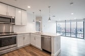 This contemporary single-floor unit is designed with a clean, modern aesthetic throughout. The heart of the space is a well-appointed kitchen featuring crisp white shaker cabinetry, light quartz countertops, and a full suite of stainless steel appliances, including a French door refrigerator, electric range, built-in microwave, and dishwasher. A peninsula with a double undermount sink serves as both a functional workspace and a natural divider between the kitchen and the open living area. Three stylish industrial-style pendant lights hang above, adding character and focused illumination.

The living area flows seamlessly from the kitchen, anchored by durable wood-look plank flooring that runs consistently throughout the space. Floor-to-ceiling windows and large black-framed glass panels flood the interior with abundant natural light while offering open views of the surrounding urban streetscape. A dedicated bedroom area features a generous triple-pane window with roller shades, providing both natural light and privacy. The neutral light grey walls and clean white trim complement the overall bright, minimalist palette. In-unit laundry is also present, adding everyday convenience. The layout's open plan and thoughtful design create a functional yet stylish living environment.