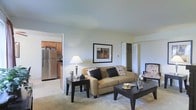 This single-floor residence offers a functional and inviting layout, anchored by a living room that serves as the central gathering space. The living area is carpeted and furnished with a tan upholstered sofa accented by decorative pillows, a rectangular coffee table, and a patterned accent chair, creating a cohesive and comfortable arrangement. A framed piece of wall art adds a decorative focal point above the seating area, while a table lamp provides warm ambient lighting.

Directly accessible from the living room, the kitchen features tile flooring, warm wood cabinetry, and stainless steel appliances including a full-sized refrigerator. The open sightline between the two spaces enhances the sense of flow and connectivity throughout the home.

A doorway visible from the living room leads to additional areas of the residence, with what appears to be a closet featuring bifold doors nearby, offering convenient storage. The neutral color palette of soft whites and beiges used throughout creates a clean, welcoming ambiance. Overall, the space is well-organized and designed to maximize practicality while maintaining a comfortable, livable atmosphere.