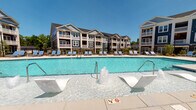 Located at 12525 Oldehurst Place in Charlotte, NC, this single-story amenity space serves as a central hub for a multi-building residential community. The exterior architecture reflects a coastal-inspired design, characterized by navy blue and gray siding, white trim accents, and stone detailing across the surrounding buildings.

The outdoor environment is a standout feature of this property. A generously sized resort-style swimming pool with crystal-clear blue water anchors the community's outdoor space, complete with a decorative fountain feature and an extensive sun deck lined with lounge chairs. The pool area is enclosed by black metal fencing and is accessible via a landscaped pathway featuring ornamental plantings and mulched garden beds.

Adjacent to the main entry is a spacious covered patio finished with stamped concrete tile flooring, providing a durable and visually appealing surface. This sheltered area is furnished with rounded modern lounge chairs and side tables, creating a comfortable outdoor seating space with open views of the surrounding greenery and pool area.

Large glass-panel entry doors and floor-to-ceiling windows allow natural light to flow freely into the interior while maintaining visual connectivity with the outdoor grounds. The landscaping throughout the community is well-maintained, incorporating a mix of shrubs, trees, and open lawn areas that contribute to a polished and welcoming atmosphere.