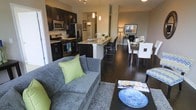 This single-floor residence showcases a thoughtfully designed open-concept layout that connects the kitchen, dining, and living areas into one cohesive, flowing space. The kitchen is outfitted with dark espresso cabinetry, stainless steel appliances including a gas range and French door refrigerator, and light-toned granite countertops that provide a striking contrast. A central island with bar seating serves as both a functional workspace and a natural dividing point between the kitchen and the rest of the living area. Pendant lighting above the island adds a refined touch, complementing the track lighting overhead. The dining area accommodates a full table with seating for multiple guests, positioned adjacent to the kitchen for easy entertaining. The living area features rich dark hardwood-style flooring that runs throughout, anchoring a comfortable seating arrangement complete with a plush sofa, area rug, and coffee table. Neutral wall tones throughout the space create a warm, inviting ambiance, while the open layout maximizes the sense of spaciousness. The overall design balances contemporary aesthetics with functional everyday living.