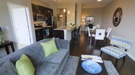 This single-floor residence showcases a thoughtfully designed open-concept layout that connects the kitchen, dining, and living areas into one cohesive, flowing space. The kitchen is outfitted with dark espresso cabinetry, stainless steel appliances including a gas range and French door refrigerator, and light-toned granite countertops that provide a striking contrast. A central island with bar seating serves as both a functional workspace and a natural dividing point between the kitchen and the rest of the living area. Pendant lighting above the island adds a refined touch, complementing the track lighting overhead. The dining area accommodates a full table with seating for multiple guests, positioned adjacent to the kitchen for easy entertaining. The living area features rich dark hardwood-style flooring that runs throughout, anchoring a comfortable seating arrangement complete with a plush sofa, area rug, and coffee table. Neutral wall tones throughout the space create a warm, inviting ambiance, while the open layout maximizes the sense of spaciousness. The overall design balances contemporary aesthetics with functional everyday living.