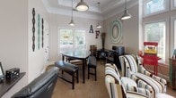Located along Primrose Parkway in Johns Creek, Georgia, this single-floor community space combines functionality with a lively, eclectic aesthetic. The interior is finished with neutral warm-toned walls, crown molding, and tray ceiling details that lend an architectural elegance to the room. Patterned carpeting in earthy tones runs throughout, anchoring the space with warmth and texture.

Industrial-style pendant lights hang from the ceiling, casting a welcoming glow over distinct functional zones. A dedicated business center area features a computer workstation with an ergonomic chair and printer, making it well-suited for productivity needs. Adjacent to this is a conference or meeting area with a dark wood table and seating, ideal for small gatherings or collaborative work.

A lounge area is furnished with bold, multi-striped wingback chairs that serve as a visual focal point, complemented by eclectic décor including decorative wall art, twig arrangements, and whimsical signage. A rustic, colorfully painted sideboard holds a coffee and beverage station, adding a hospitality-forward touch.

Large windows along one wall allow natural light to filter in, offering views of the lush outdoor surroundings. A standout feature is a classic red popcorn machine, adding a playful and social element to the space. Overall, the area balances comfort, practicality, and personality in a distinctive community setting.