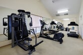 Located at 300 Hilary Way in Vallejo, California, this fitness center offers a well-organized, single-floor layout designed to support a full range of workout routines. The space is finished with light-colored carpet flooring and warm, cream-toned walls, creating a clean and inviting environment for exercise.

The room is illuminated by overhead fluorescent lighting and benefits from natural light streaming through multiple slatted windows. Full-length mirrors are mounted along one wall, amplifying the sense of space and providing visual feedback during workouts.

The fitness center is well-stocked with cardio equipment, including multiple treadmills, elliptical trainers, and a stationary rowing machine. Strength and flexibility training is supported by a cable resistance machine, step platforms, resistance bands, exercise mats, and stability balls — including a BOSU balance trainer. A wall-mounted television provides entertainment during workouts, and a standing fan adds to user comfort.

Decorative indoor plants and signage contribute to a welcoming atmosphere. A glazed door on one end provides access to an adjacent outdoor area, adding a sense of connectivity to the surrounding environment. The space is thoughtfully arranged to allow ease of movement between equipment, making it a functional and practical amenity for residents at this Vallejo location.