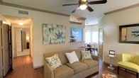 This single-floor residence offers a thoughtfully designed open-concept layout that seamlessly connects the living, dining, and kitchen areas into one cohesive living space. The living area is anchored by warm hardwood flooring that extends throughout, creating a sense of continuity and warmth. A ceiling fan with integrated lighting adds both comfort and charm, while classic crown molding along the ceiling lends a refined, traditional character to the space.

The living room provides a welcoming atmosphere, with ample wall space currently showcasing large-scale artwork that adds a vibrant, artistic focal point. A pass-through opening with a small shelf connects the living area to the kitchen and dining space, allowing for easy interaction between rooms and enhancing the open feel of the layout.

The dining area benefits from natural light streaming in through a large window, creating a bright and airy environment ideal for everyday meals or entertaining. A pendant light fixture above the dining space adds a decorative touch. The kitchen features a bar-height counter, perfect for casual dining or serving. The front entry is highlighted by a bold accent wall in a soft blue hue, adding a pop of color and a welcoming character to the home's interior.