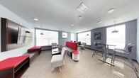 This single-floor common lounge is designed to accommodate a variety of activities, from casual socializing to focused work sessions. The space is finished with neutral carpeting and gray-painted walls, creating a clean, contemporary backdrop that is accented throughout by bold red furnishings. Multiple large windows line the perimeter, allowing abundant natural light to fill the room while offering views of the surrounding exterior. Pendant lights with industrial-style shades hang from the textured ceiling, supplementing the natural light and contributing to the modern aesthetic. The layout is thoughtfully organized into distinct seating zones. A central area features a low work table paired with a cushioned accent chair and a round, upholstered ottoman in red and white. A prominent red-backed banquette sofa serves as a visual centerpiece, flanked by bar-height tables and seating for more elevated, collaborative use. Along the perimeter, window ledges with built-in bench seating in red provide additional spots for relaxation. A tall bar table with stools near the far wall offers yet another option for informal gathering or individual work, making this lounge a well-rounded, multi-use communal space.