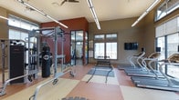 Located in Littleton, Colorado, this single-floor fitness center at 13195 West Progress Avenue is a comprehensively equipped workout facility designed to support a wide range of exercise routines. The space features a vibrant, energetic atmosphere accented by bold red and neutral taupe walls, complemented by a durable, multi-toned rubber flooring ideal for high-intensity training.

The main workout area is outfitted with treadmills and cardio equipment along one wall, while a dumbbell rack with a full range of free weights provides ample options for strength training. A dedicated functional training zone features a heavy punching bag, a multi-use power rack, and battle ropes, offering versatility for combat-style and cross-training workouts.

A glass-partitioned room within the facility provides a separate area for cycling or additional equipment, maintaining visual openness while defining distinct workout zones. Natural light flows in through large windows, offering views of the surrounding Colorado landscape, and a wall-mounted flat-screen TV adds an entertainment element to cardio sessions.

High ceilings with industrial-style linear lighting fixtures and a ceiling fan ensure the space feels open and well-ventilated. The overall design balances functionality with an energizing ambiance, making this fitness center a well-rounded destination for residents and members alike.