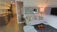 This single-floor space offers a well-appointed and cohesive interior design that balances comfort with contemporary style. The living area serves as the focal point, featuring a plush light grey sofa adorned with colorful patterned and solid accent pillows in shades of green and floral prints. The walls are finished in a two-tone palette — a soft sage green upper section complemented by a clean white lower half — creating a calm and inviting atmosphere. A curated arrangement of circular mirrors in varying sizes decorates the feature wall, adding depth and a decorative focal point to the room. Warm ambient lighting from a tall floor lamp enhances the cozy feel of the space. The flooring throughout is a light wood-tone material that adds warmth and continuity. A glass-paneled partition wall separates the living area from an adjacent corridor or entryway, allowing natural light to pass through while maintaining a sense of openness. Accent furniture, including a minimalist black side table and a round red accent table, add contrast and personality. The overall ambiance is modern, relaxed, and carefully curated, making it a comfortable and visually engaging space.