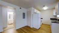 This single-floor space features a clean, functional layout with a bright and minimalist aesthetic. The kitchen area is outfitted with white appliances, including a full-sized refrigerator and a range, complemented by white upper cabinetry that provides ample storage. The light hardwood flooring runs throughout, lending warmth and continuity to the space. Crisp white walls enhance the sense of openness and allow natural and artificial light to reflect effectively throughout the interior. A louvered closet door offers additional storage, while a visible electrical panel is conveniently located along one wall. Ceiling-mounted flush lighting provides consistent illumination, and HVAC vents are positioned in both the ceiling and walls to ensure comfortable climate control. The open layout between the kitchen and adjacent areas creates an unobstructed flow, making the space feel connected and easy to navigate. The overall design is straightforward and practical, with a neutral palette that provides a versatile foundation suitable for a variety of furnishing styles and uses.