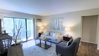This single-floor space offers a comfortable and thoughtfully designed interior that blends modern style with a warm, inviting ambiance. The living room serves as the centerpiece of the space, bathed in natural light that streams through large sliding glass doors leading to a private balcony. The balcony overlooks a serene, tree-lined setting, bringing a sense of nature and tranquility directly into the home. The living room is well-proportioned, providing ample space for both relaxed lounging and casual entertaining. Neutral wall tones and light-colored flooring create a clean, airy backdrop that complements the contemporary furnishings. A wall-mounted air conditioning unit ensures year-round comfort, while tasteful décor — including a large nature-inspired wall art piece — adds a refined, cohesive aesthetic. The space is illuminated by soft ambient table lamps that enhance the room's warm and relaxed atmosphere. Overall, this space combines functional design with natural surroundings, making it an ideal retreat that balances indoor comfort with scenic outdoor connectivity.