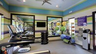 This fitness center makes a bold visual statement with its eye-catching horizontally striped walls in shades of blue, green, purple, and gray, creating an energetic and motivating atmosphere. Large mirrors with dark frames line the walls, visually expanding the space and allowing users to monitor their form during workouts. A ceiling fan is centrally mounted overhead, complementing the recessed lighting and a wall-mounted air conditioning unit to ensure comfortable airflow throughout sessions. The space is well-equipped with a row of cardio machines, including treadmills, visible along one wall. A dumbbell rack stocked with a full range of free weights sits prominently on the floor, accompanied by a weight bench. Along the opposite side, a dedicated stretching and functional fitness area features an organized rack holding a colorful assortment of stability balls in yellow, green, and orange, along with medicine balls and foam rollers. A flat-screen television is mounted on the mirrored wall, providing entertainment during workouts. Natural light filters in through windows, contributing to the bright and welcoming environment of this comprehensively outfitted fitness facility.