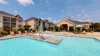This outdoor pool area is designed to deliver a resort-like recreational experience. The centerpiece is a generously sized swimming pool with clear, blue water, tiled edges, and a built-in in-pool basketball hoop for active recreation. A wide, open concrete sundeck surrounds the pool and is furnished with multiple lounge chairs and shade umbrellas, providing ample space for relaxing under the open sky.

Adjacent to the pool is a covered cabana structure that serves as a shaded retreat. It features distinctive turquoise shiplap walls, a white beadboard ceiling, and stacked-stone base columns that lend a polished, coastal aesthetic. Inside the cabana, a stone-faced bar counter with a granite top is lined with wicker bar stools, creating a comfortable gathering spot. A wall-mounted flat-screen television and an arched window overlooking the community complete the space. Wicker seating and a round glass-top table provide additional lounging options within the covered area.

The entire pool complex is set against a backdrop of multi-story craftsman-style residential buildings with pitched rooflines, dormer windows, and landscaped greenery. A separate clubhouse pavilion with a prominent arched entryway and stone detailing is visible nearby, reinforcing the well-appointed character of the overall community amenity space.
