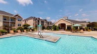 This outdoor pool area is designed to deliver a resort-like recreational experience. The centerpiece is a generously sized swimming pool with clear, blue water, tiled edges, and a built-in in-pool basketball hoop for active recreation. A wide, open concrete sundeck surrounds the pool and is furnished with multiple lounge chairs and shade umbrellas, providing ample space for relaxing under the open sky.

Adjacent to the pool is a covered cabana structure that serves as a shaded retreat. It features distinctive turquoise shiplap walls, a white beadboard ceiling, and stacked-stone base columns that lend a polished, coastal aesthetic. Inside the cabana, a stone-faced bar counter with a granite top is lined with wicker bar stools, creating a comfortable gathering spot. A wall-mounted flat-screen television and an arched window overlooking the community complete the space. Wicker seating and a round glass-top table provide additional lounging options within the covered area.

The entire pool complex is set against a backdrop of multi-story craftsman-style residential buildings with pitched rooflines, dormer windows, and landscaped greenery. A separate clubhouse pavilion with a prominent arched entryway and stone detailing is visible nearby, reinforcing the well-appointed character of the overall community amenity space.